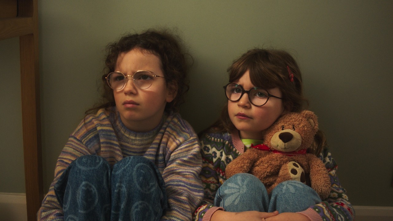 Two young sisters sitting together against a bedroom wall in the Sharps Sisters advert